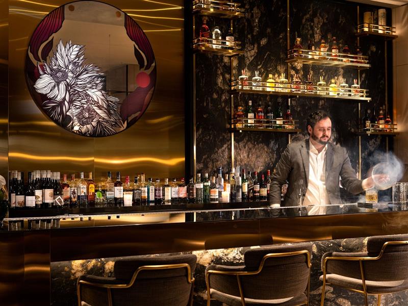 An image of a bar with low seating, bar stools, artwork, drinks on shelves and a bartender.