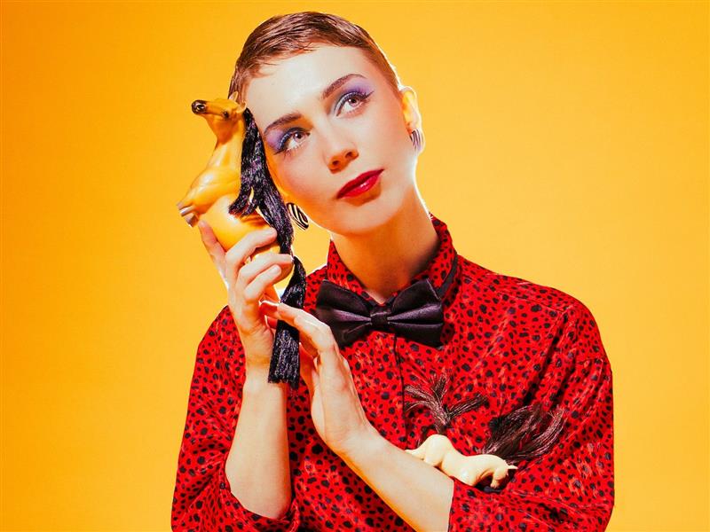 Cabaret Artist FRANKLY humorously holding a toy horse to her year, as if it were a phone.