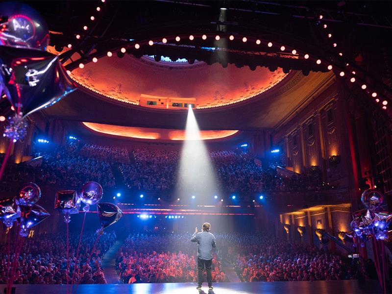 the back of a comedian, performing on stage, taken looking out from the stage to the audience