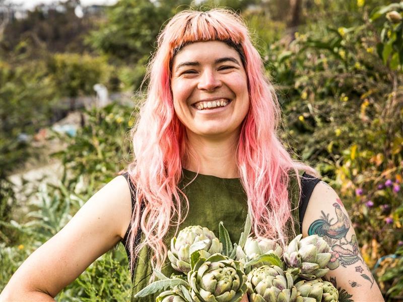 Hannah Moloney wears green and stands in front of a green bush holding an armful of artichokes